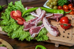 Fresh lamb chops arranged on lettuce with sliced tomatoes and a green chili pepper, surrounded by colorful vegetables and spices.