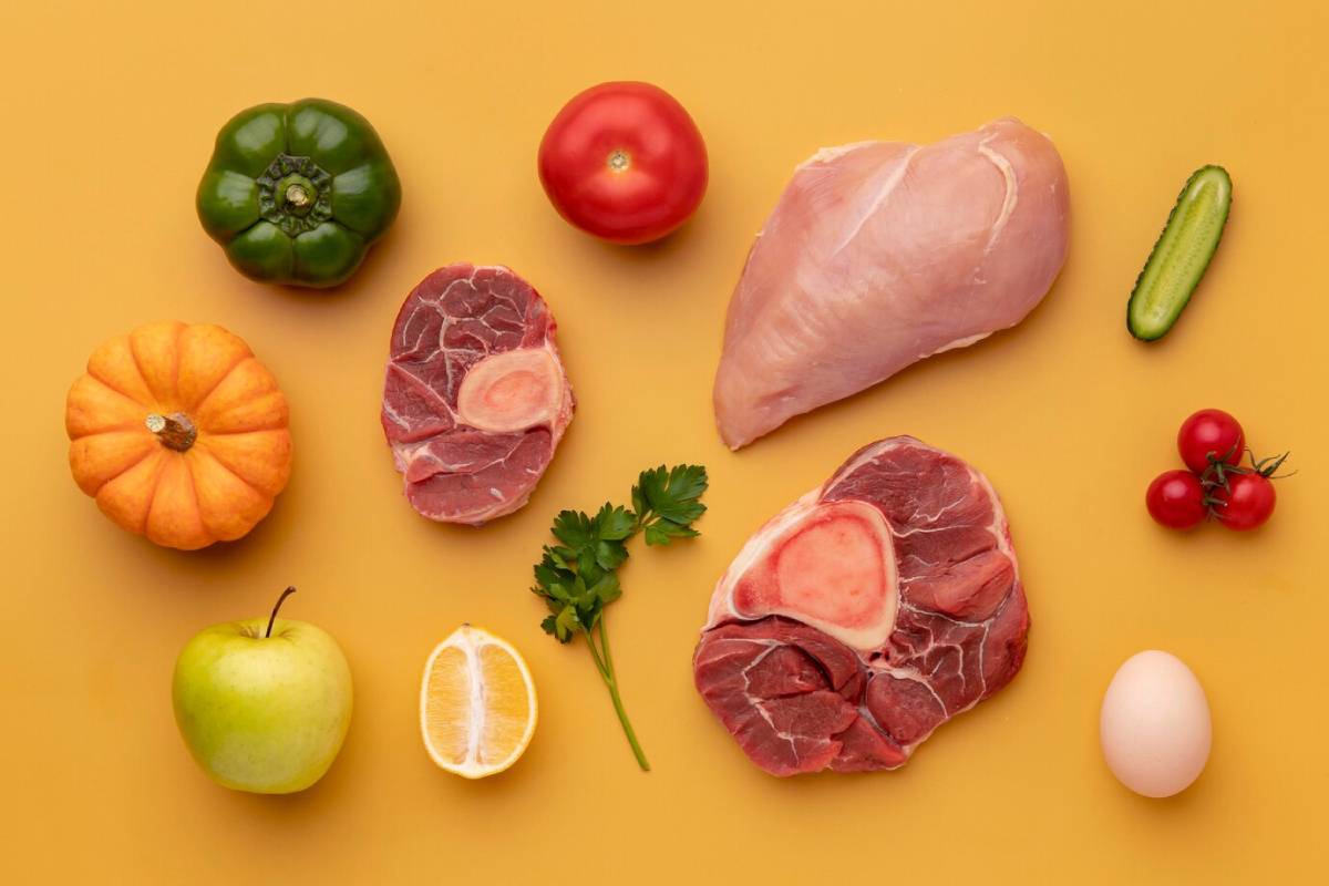 An assortment of fresh foods on a yellow background, including meats, vegetables, fruits, an egg, and herbs.