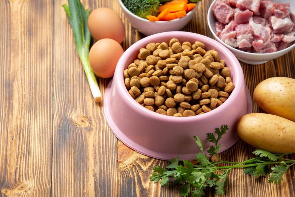 A pink bowl filled with pet food surrounded by eggs, chopped meat, vegetables, and potatoes on a wooden surface.