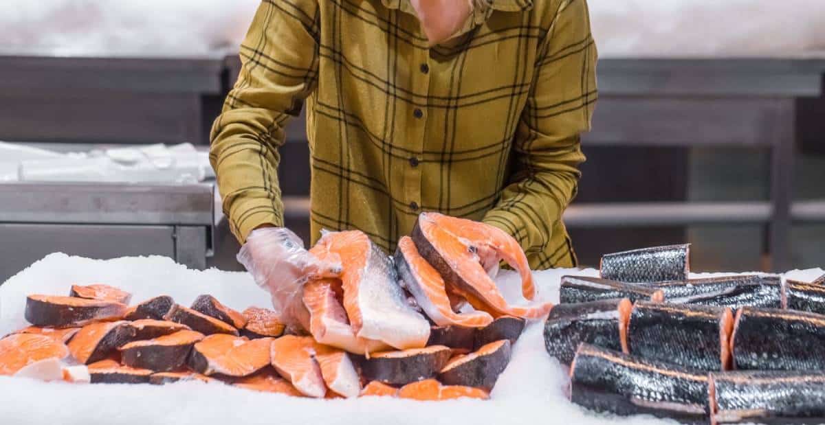A person in a plaid shirt sorts salmon fillets on a bed of ice at a seafood counter, wearing gloves for hygiene.