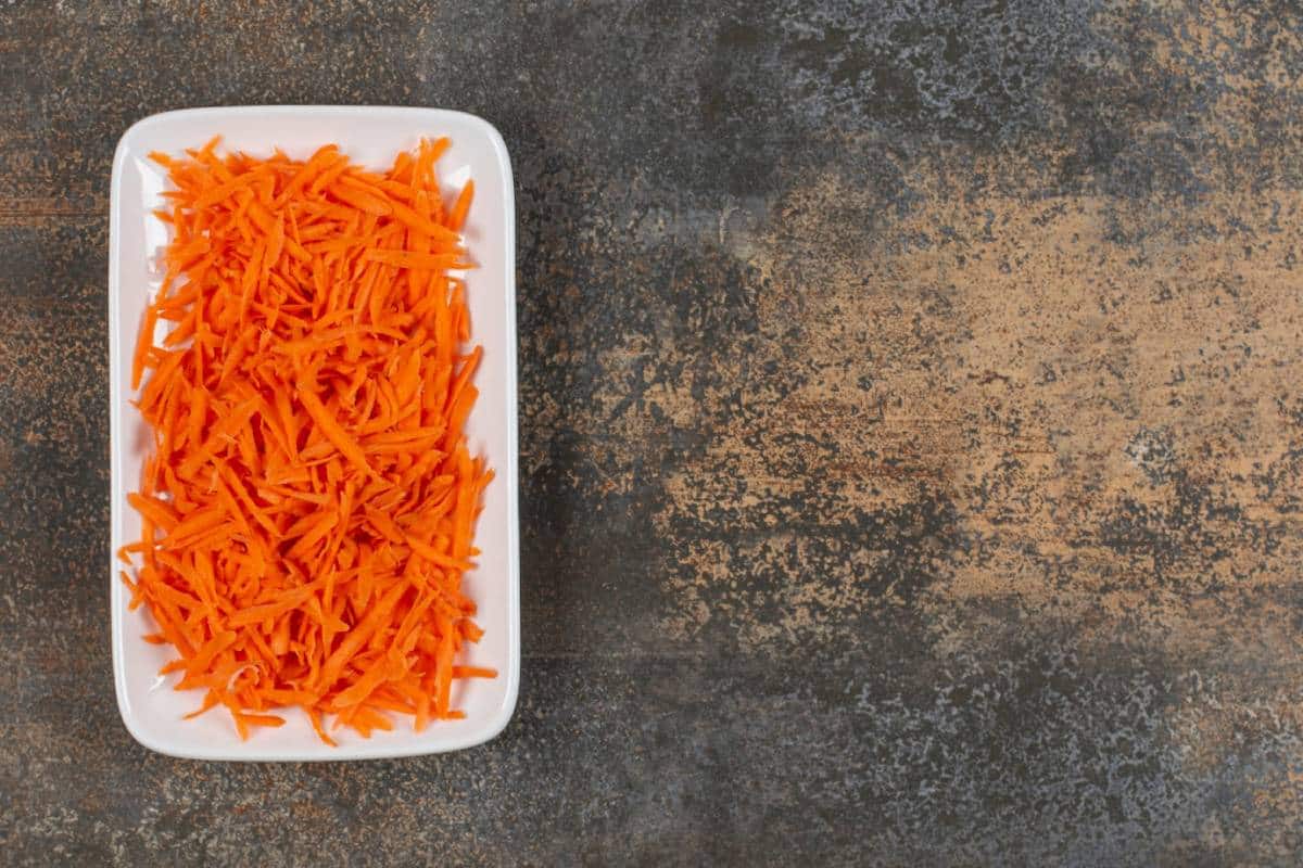 A white dish filled with finely shredded orange carrots, resting on a textured dark surface.