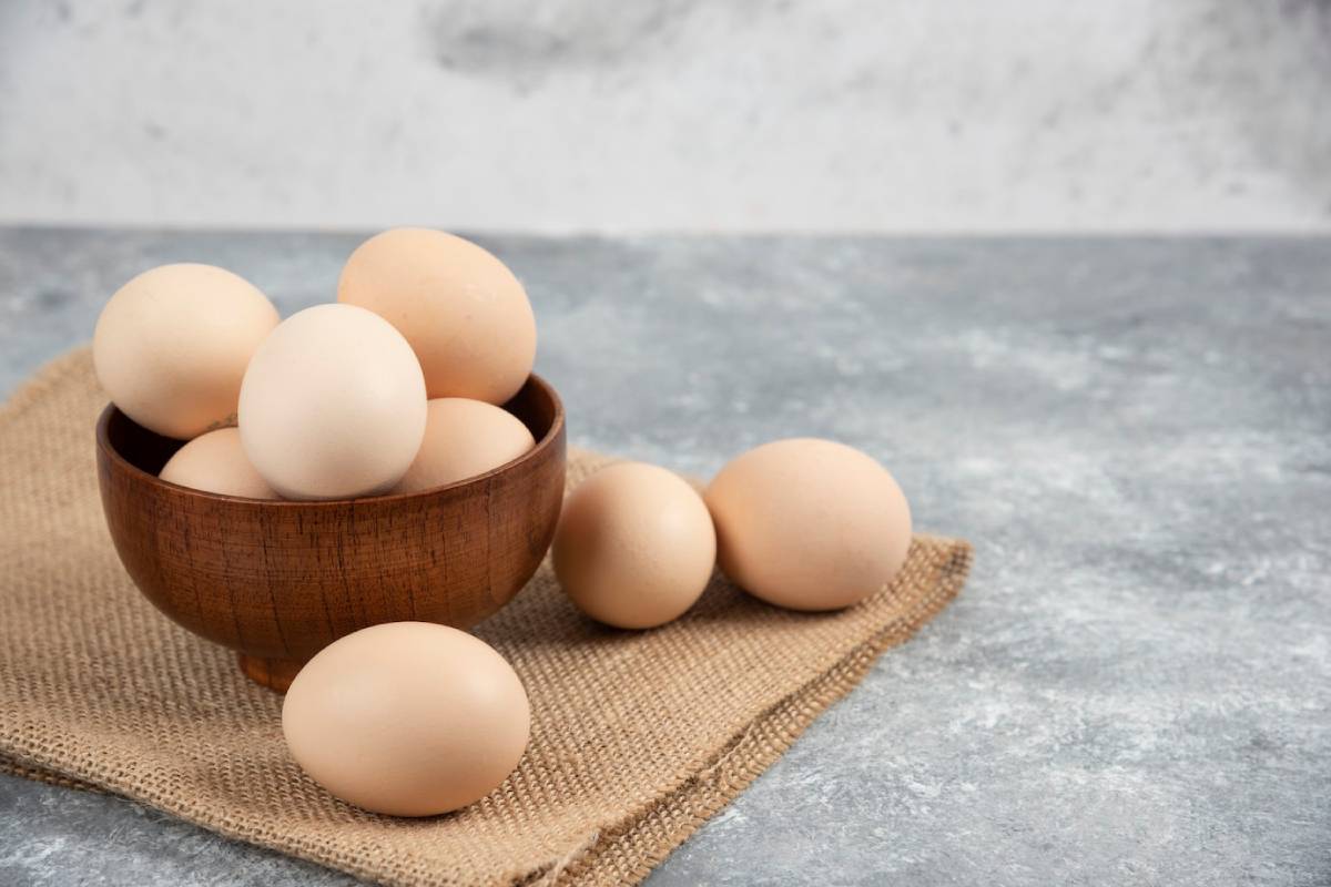 A wooden bowl filled with pale brown eggs, surrounded by a few more on a burlap cloth, set against a textured gray background.