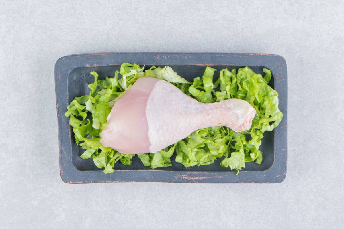 Raw chicken drumstick resting on a bed of fresh, shredded lettuce on a dark serving tray, set against a light background. 