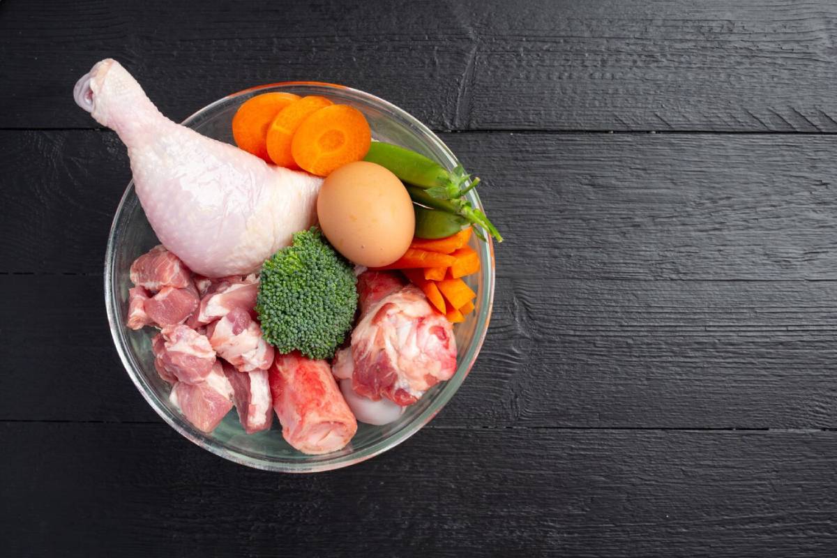 A glass bowl filled with chicken drumstick, meat cuts, an egg, broccoli, carrots, and green beans on a dark wooden surface.
