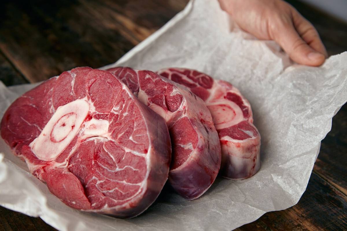 Three raw, meaty osso buco cuts, neatly arranged on parchment paper, with a hand reaching to lift them off a wooden surface.
