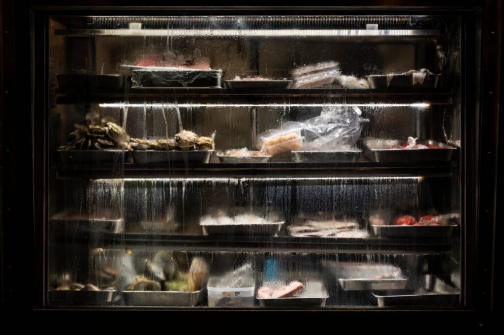 A glass-fronted refrigerator displaying various containers of seafood and meat, surrounded by condensation and water droplets.
