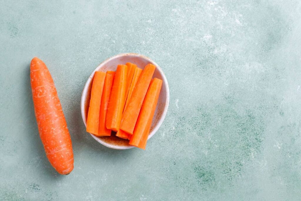 A whole carrot next to a bowl filled with sliced carrot sticks, set against a light green textured background.