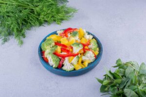 A vibrant salad featuring broccoli, red bell peppers, and yellow peppers, served in a blue bowl, surrounded by fresh herbs.