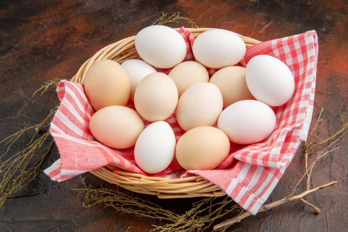 A wooden basket filled with a variety of eggs, both white and brown, resting on a red and white checkered cloth. 