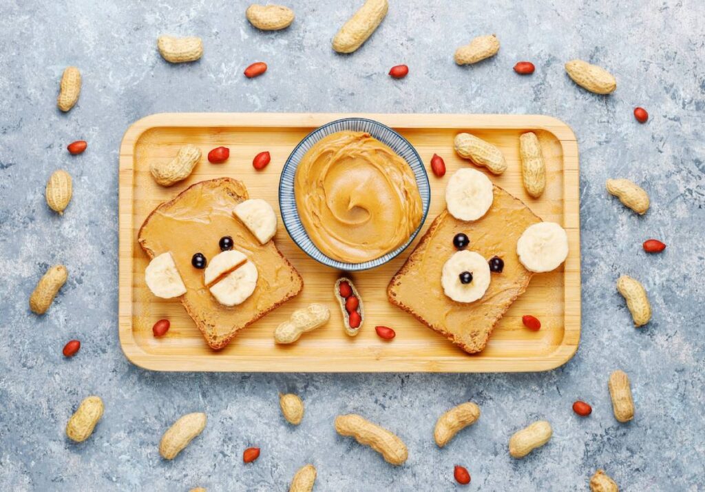 A wooden tray holds two peanut butter toast bears, decorated with bananas and blueberries, surrounded by peanuts and a bowl of peanut butter.