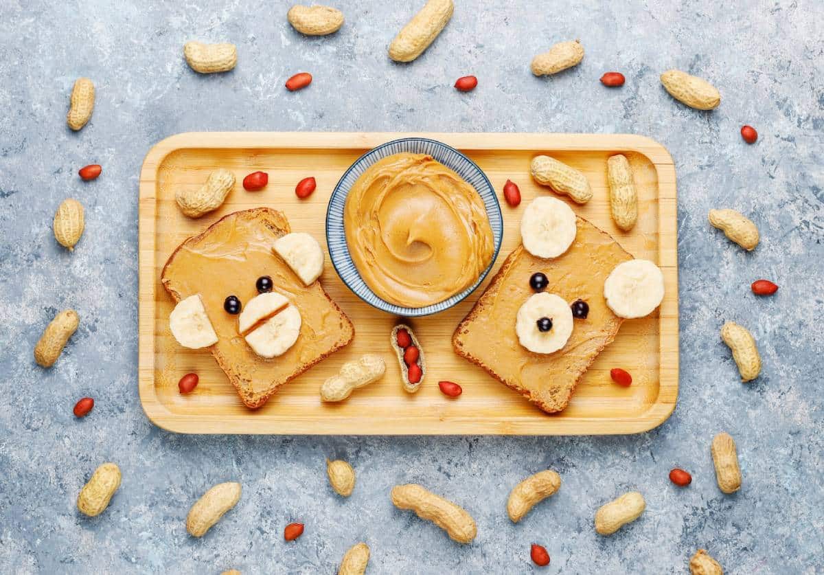 A wooden tray holds two peanut butter toast bears, decorated with bananas and blueberries, surrounded by peanuts and a bowl of peanut butter.