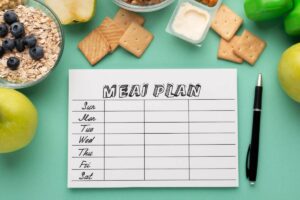 A blank meal plan sheet with days of the week, surrounded by fresh fruits, oats, crackers, and yogurt on a turquoise background.