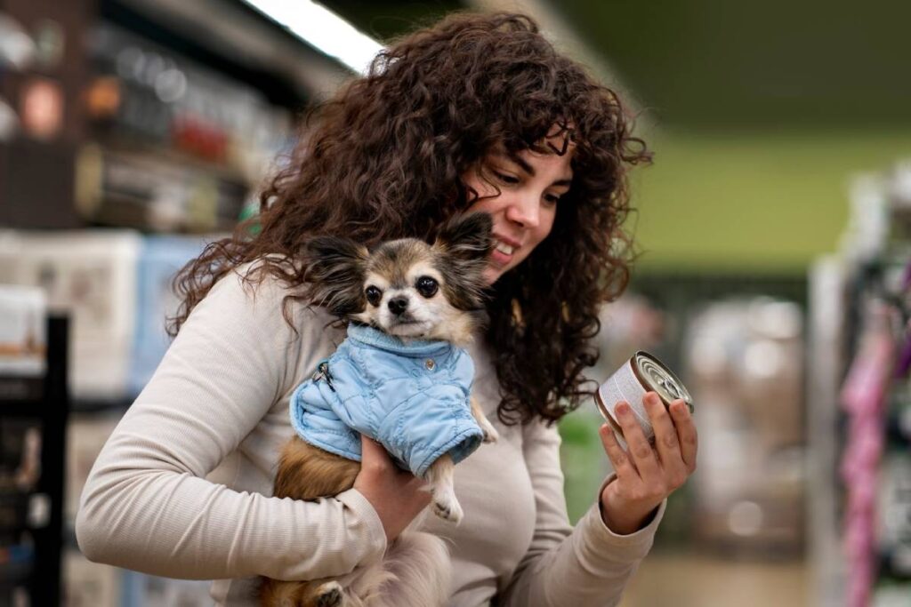 A woman holds a small dog in a blue coat while examining a can in a pet store surrounded by shelves of pet supplies.