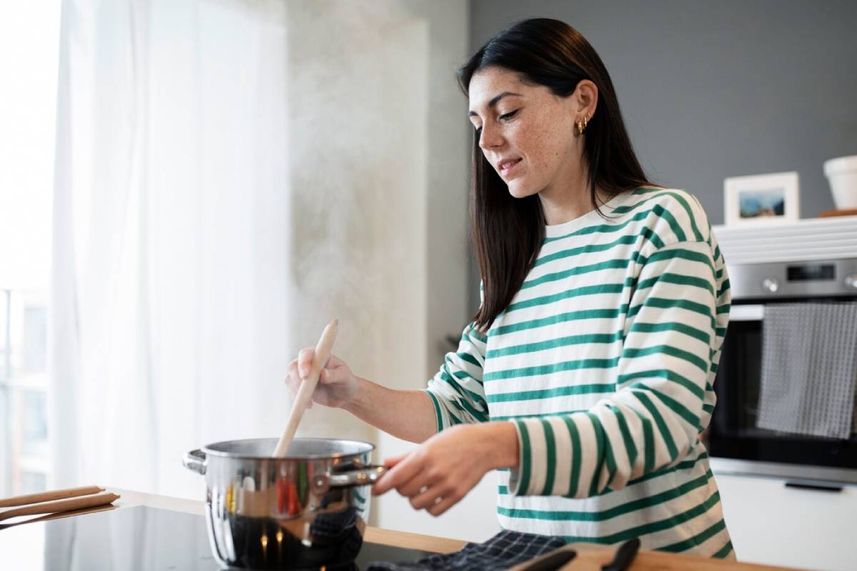 A person in a striped sweater stirs a pot on the stove, surrounded by kitchen tools and steam rising from the pot.