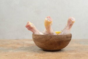Three raw chicken drumsticks seasoned with spices, placed upright in a rustic wooden bowl on a textured countertop.