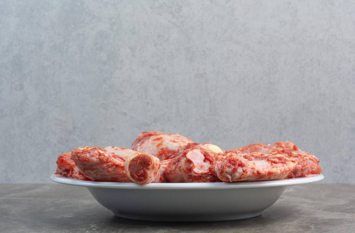 A white plate holds a large portion of raw, marbled beef bones, with a textured gray background.
