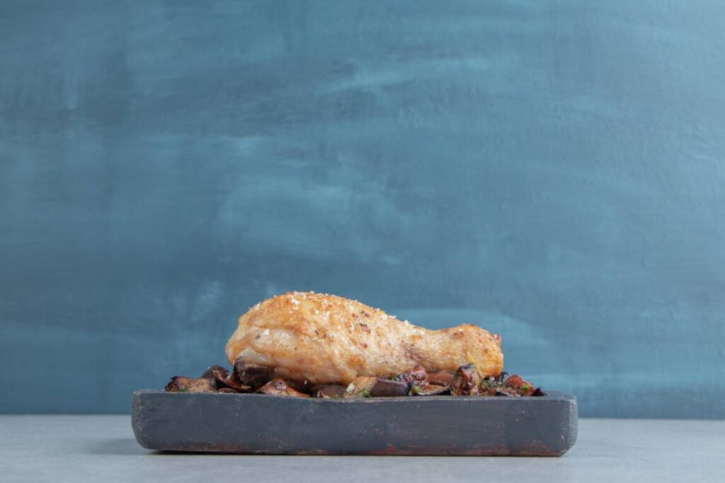 A golden-brown chicken drumstick sits atop a bed of roasted vegetables on a dark plate against a blue backdrop.