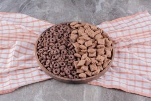 A wooden bowl filled with two types of chocolate cereal: round puffs on one side and pillow-shaped pieces on the other, on a checkered cloth.
