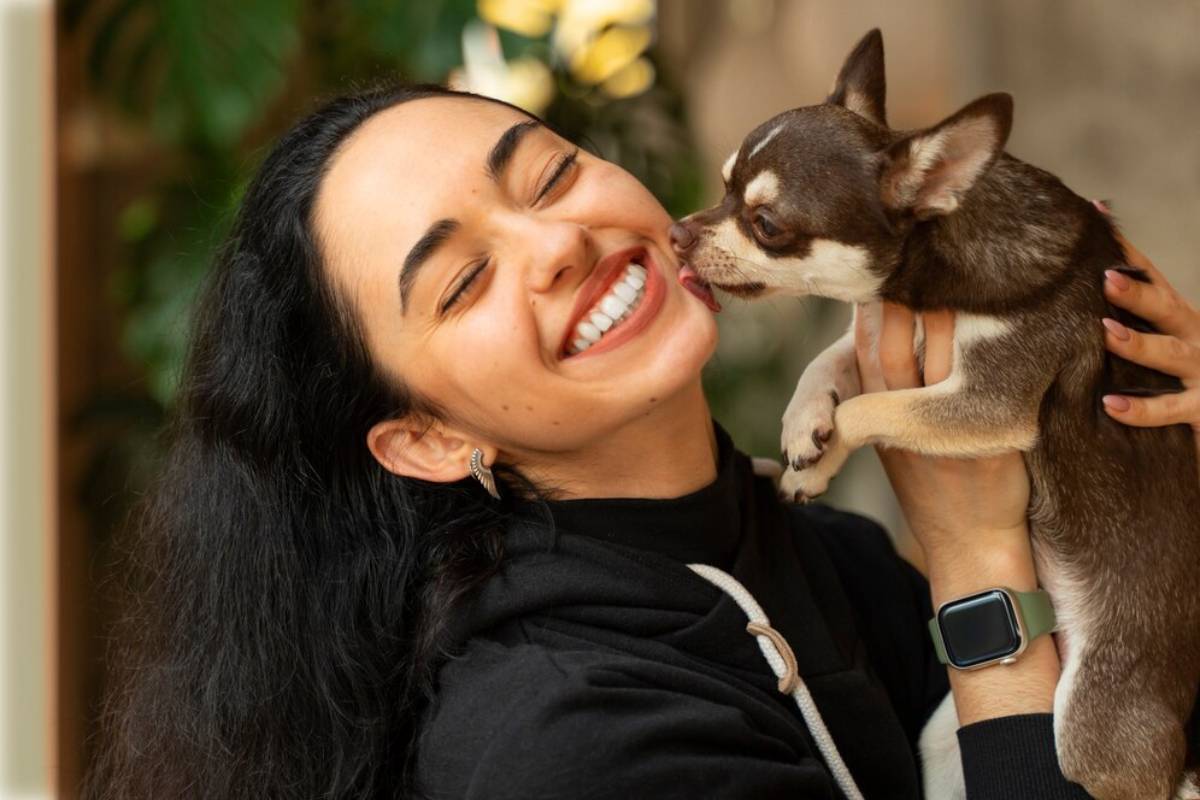 A person holds a small brown and white Chihuahua close, with greenery blurred in the background, capturing a moment of affection. 