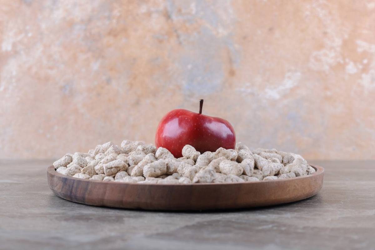A shiny red apple sits atop a mound of textured, light-colored cereal in a wooden bowl against a neutral background. 