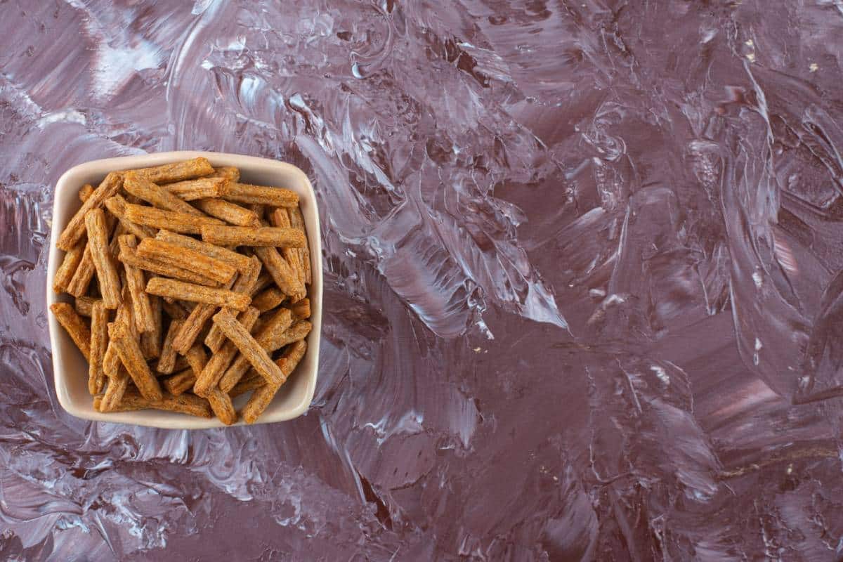 A bowl filled with crunchy, rectangular snack sticks on a textured, mauve-colored background.