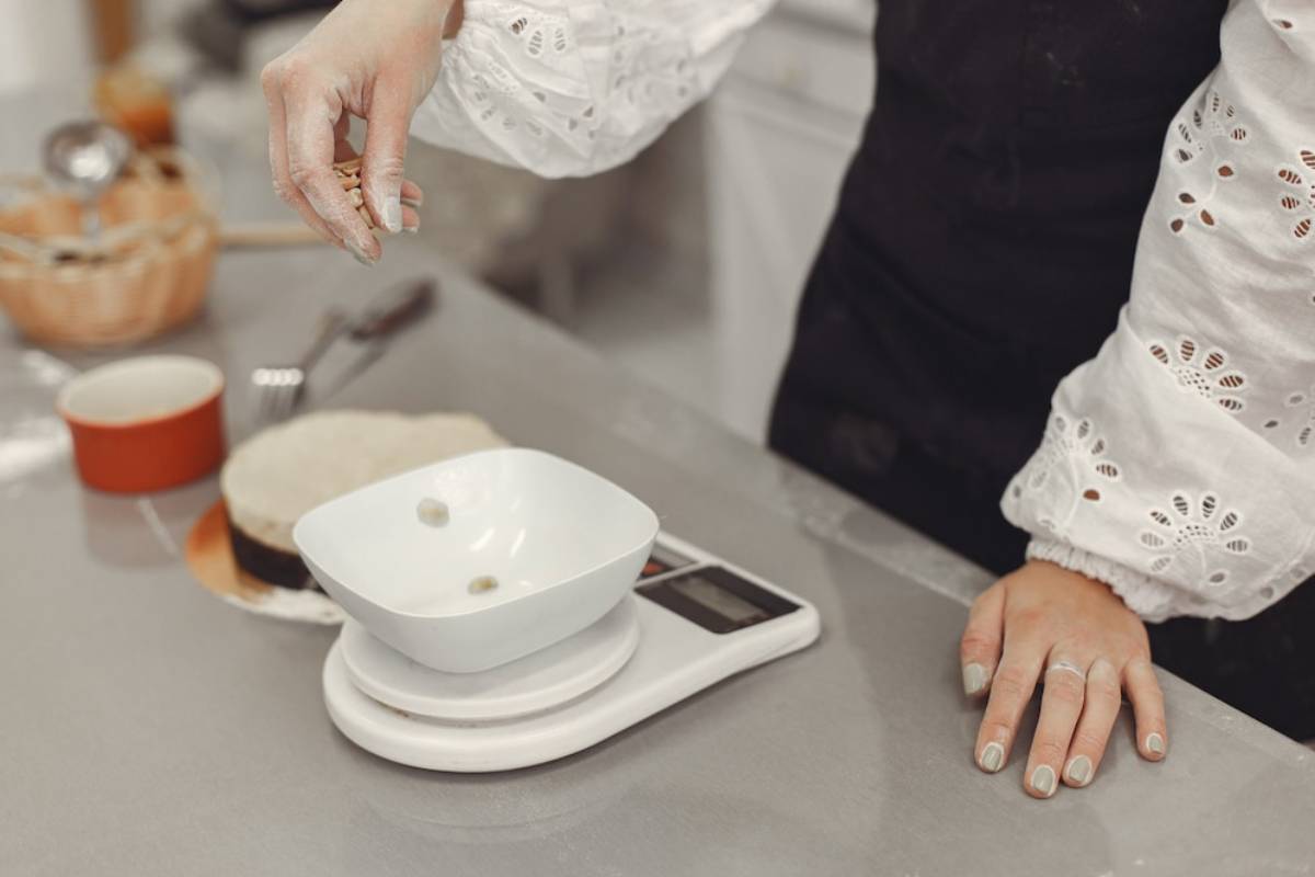 A person in a white shirt measures ingredients on a digital scale in a kitchen, with flour on their hand and countertop.