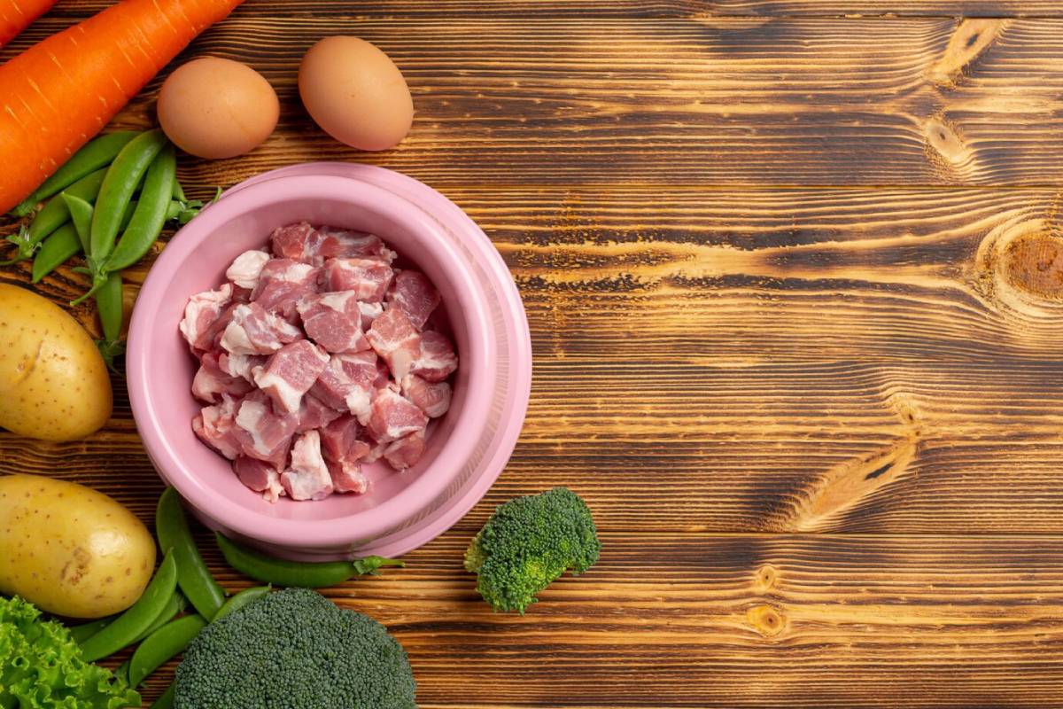 A pink bowl filled with chopped meat, surrounded by carrots, potatoes, eggs, broccolini, and snap peas on a wooden surface. 