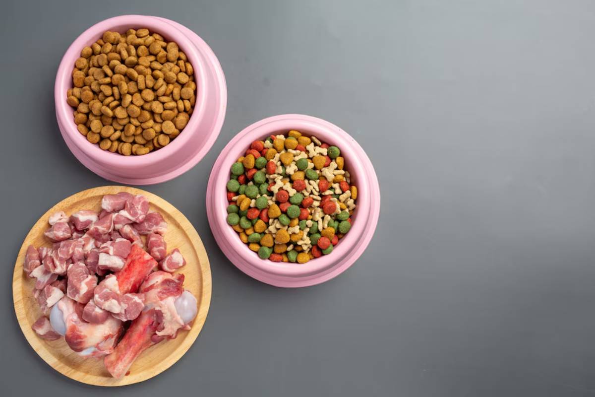 Two pink bowls filled with dry pet food and colorful kibble, alongside a wooden plate with raw meat and bones. 