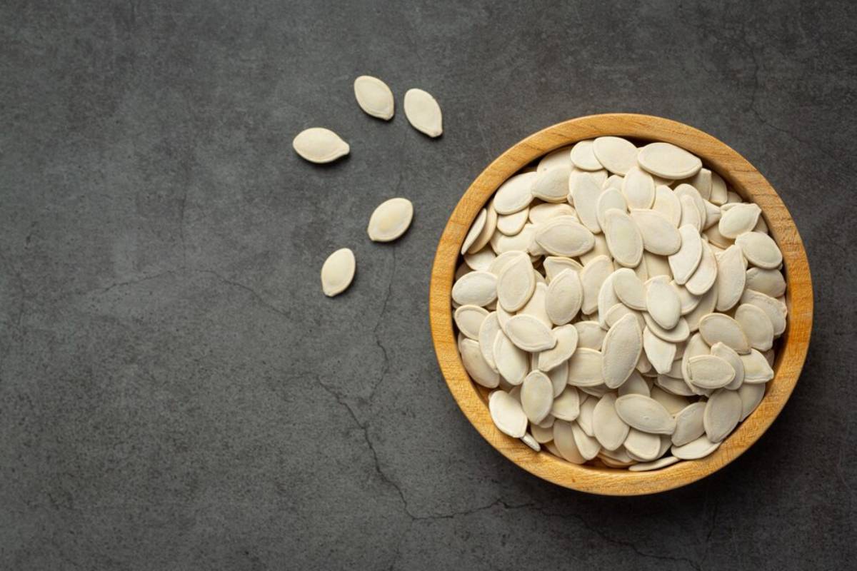 A wooden bowl filled with pumpkin seeds, with a few scattered on a dark stone surface. 