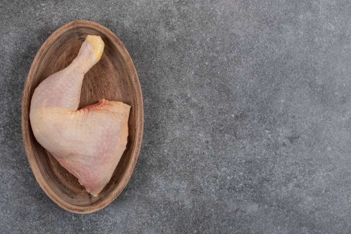 A raw chicken leg quarter placed on a wooden oval plate against a gray background.