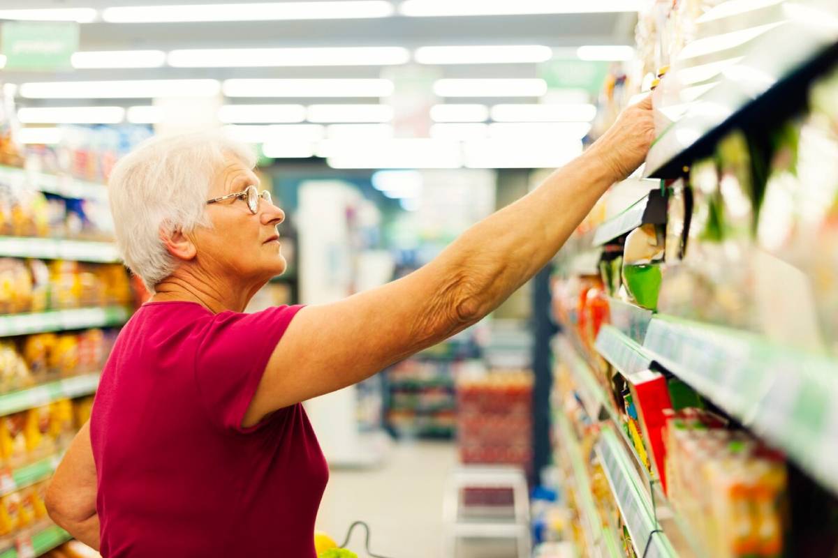 An elderly woman in a burgundy shirt reaches for products on a  store shelf, surrounded by colorful packaging. 
