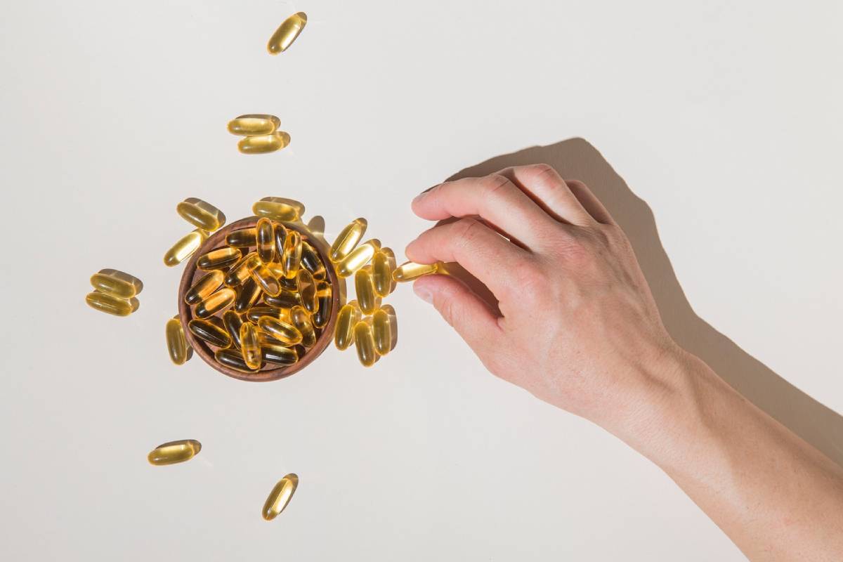 A hand reaching for dietary capsules in a wooden bowl, with many capsules scattered on a light surface. 