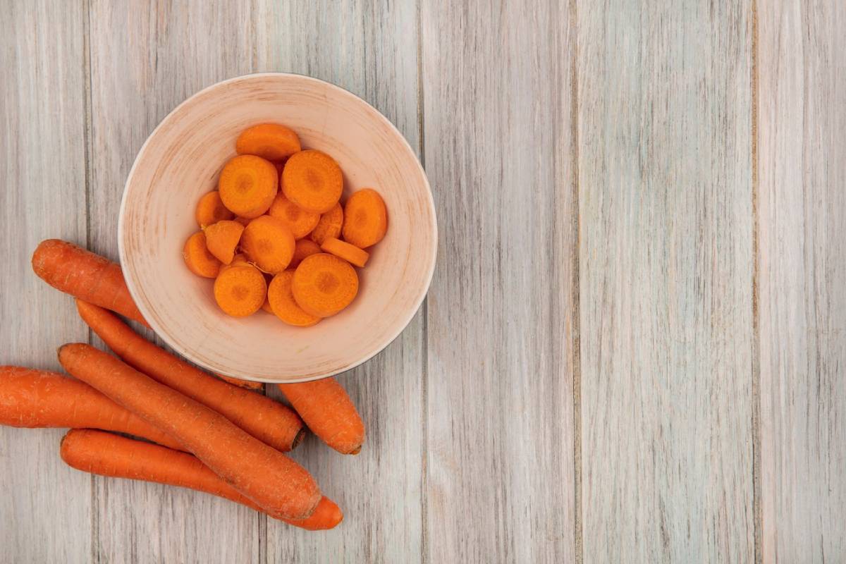 A bowl of bright orange carrot slices sits atop a wooden surface, beside whole carrots scattered around. 