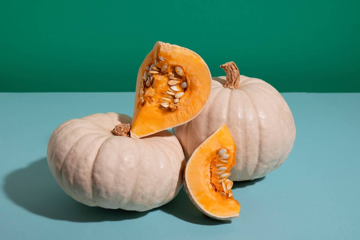 Three pale pumpkins sit on a blue surface, with one sliced open to reveal its vibrant orange flesh and seeds, against a green backdrop.