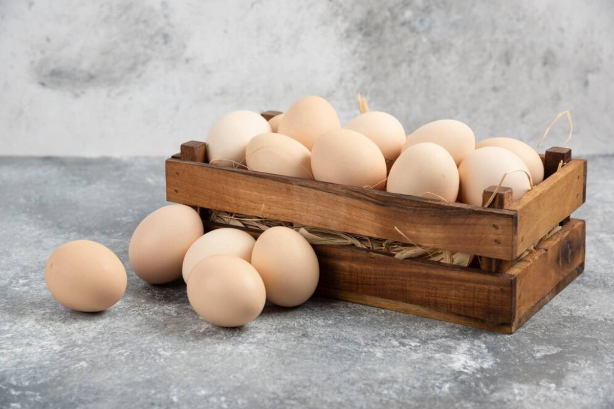 A wooden crate filled with fresh, light brown eggs, with some eggs scattered on a textured gray surface.